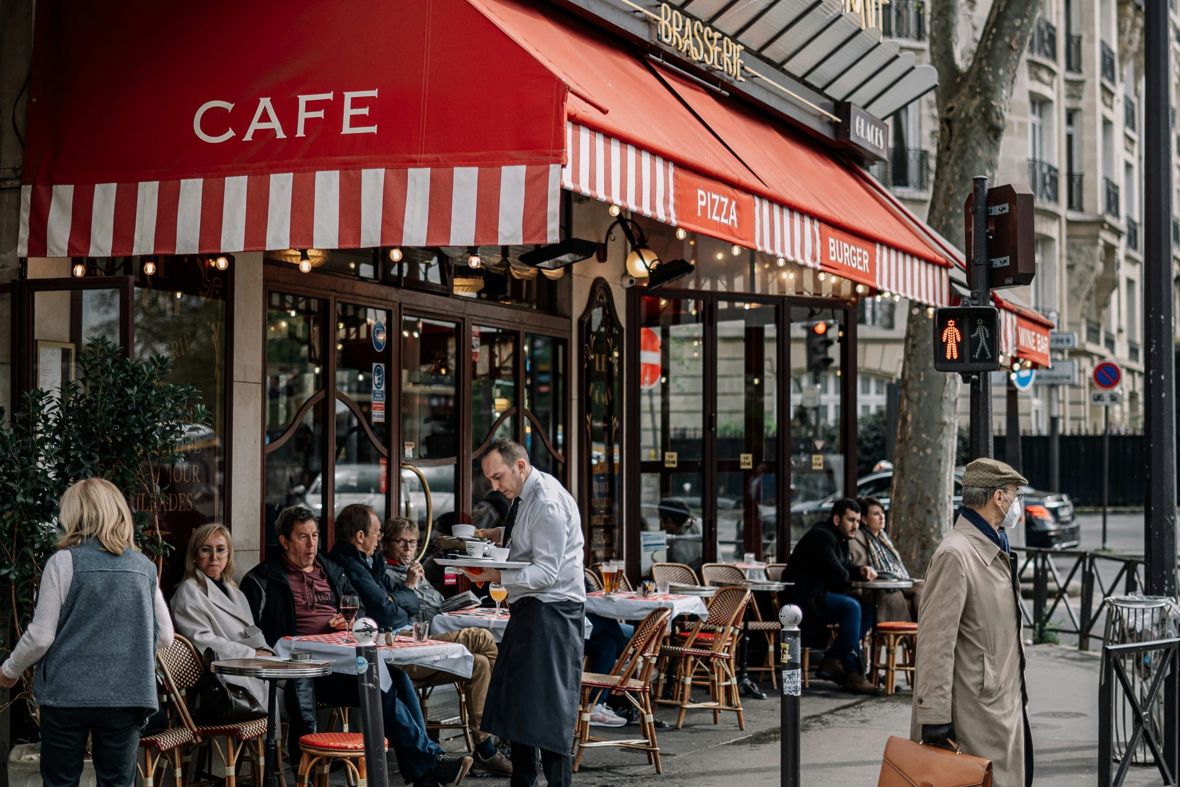 よその国の、角にある喫茶店の外観。赤い日除けに CAFE と白抜きで入っていて、歩道に丸いテーブルと籐の椅子が並び、客が座っている。給仕がカップを載せた盆を運んでいる。